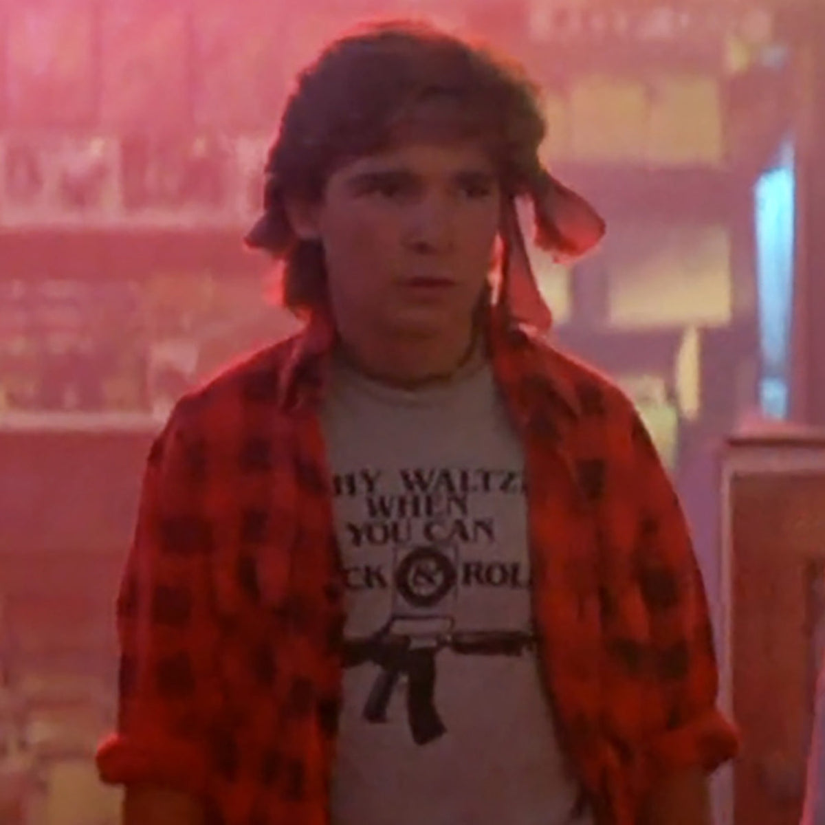Corey Feldman as Edgar Frog in The Lost Boys, wearing a red checked shirt over a grey “Why Waltz When You Can Rock & Roll” T-shirt.