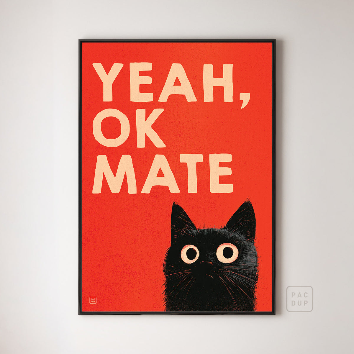 Art print with cream “Yeah, OK Mate” lettering and a wide-eyed black cat on red, displayed in a black frame.
